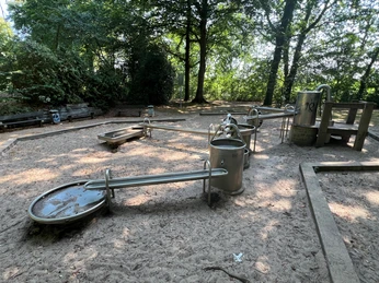 Wasserspielplatz mit Sandboden, großer Baumbestand, mehrere Edelstahlspielelemente für Kinder.