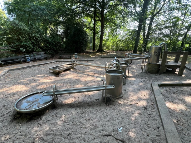 Wasserspielplatz mit Sandboden, großer Baumbestand, mehrere Edelstahlspielelemente für Kinder.