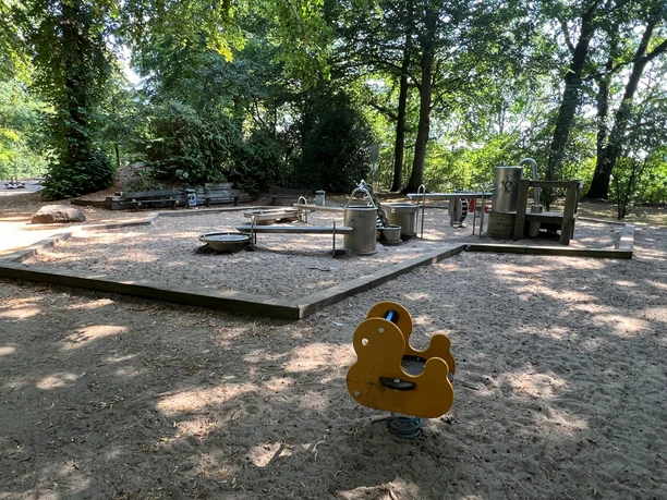 Abenteuerlicher Wasserspielplatz im Wald, Metallsprinkler und Sand, umgeben von hohen Bäumen.