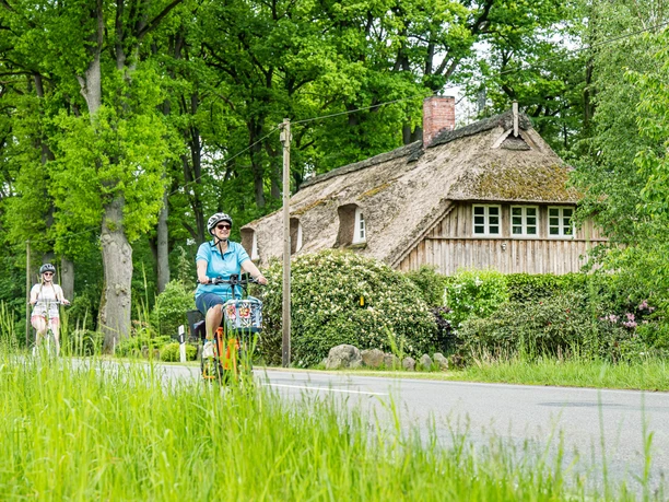 Drei Frauen radeln an einem reetgedeckten Bauernhaus unter grünen Bäumen vorbei.