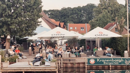 Kranenborgh - Blick auf den Steg Personen entspannen sich im Kranenborgh Biergarten am Wasser unter schattigen Sonnenschirmen.