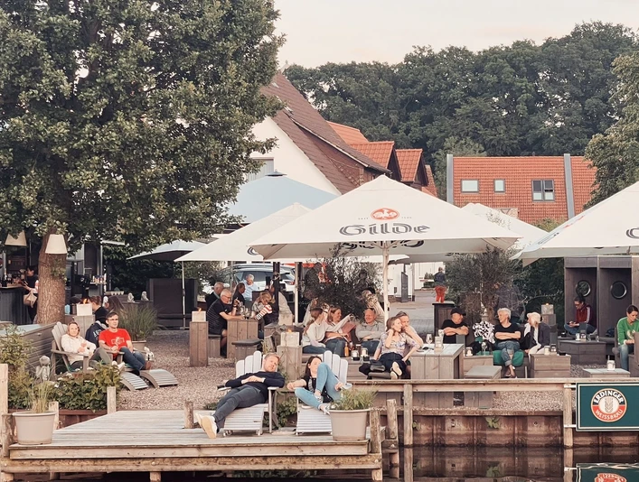 Kranenborgh - Blick auf den Steg Personen entspannen sich im Kranenborgh Biergarten am Wasser unter schattigen Sonnenschirmen.