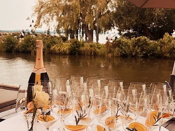 Kranenborgh - Getränke Ein gedeckter Tisch mit Sektflasche, Gläsern mit Orangen und Rosmarin vor einem Fluss im Freien.