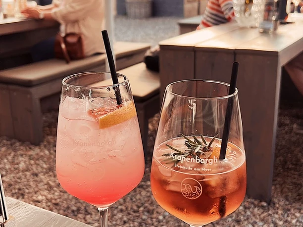 Kranenborgh Biergarten Zwei farbenfrohe Cocktails mit Strohhalmen auf einem Tisch, Gäste im Hintergrund an langen Holzbänken.