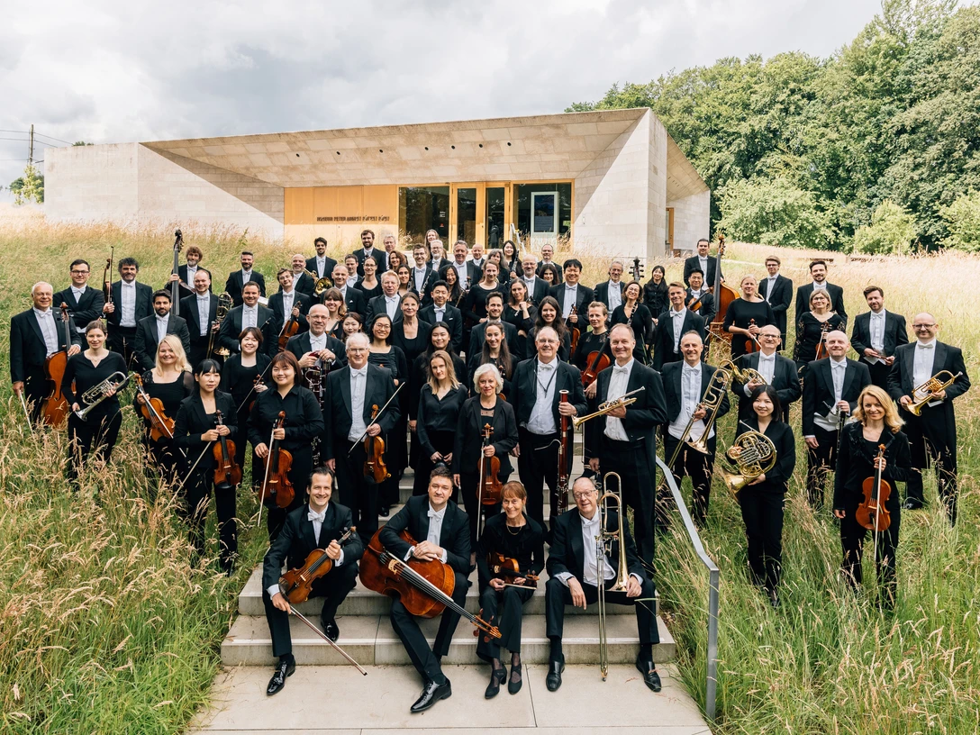 NWD_Orchesterfoto_Copyright Michael Adamski.jpg Eine große Orchestergruppe posiert in hellgrüner, natürlicher Umgebung vor einem modernen Gebäude.