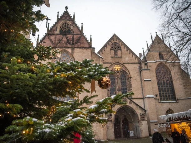 Weihnachtsmarkt mit beleuchtetem Tannenbaum und gotischer Kirche im Hintergrund, Schnee fällt sanft.