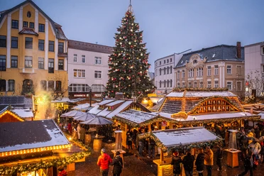 Weihnachtsmarkt mit leuchtendem Tannenbaum, geschmückten Ständen und Menschen in winterlicher Atmosphäre.