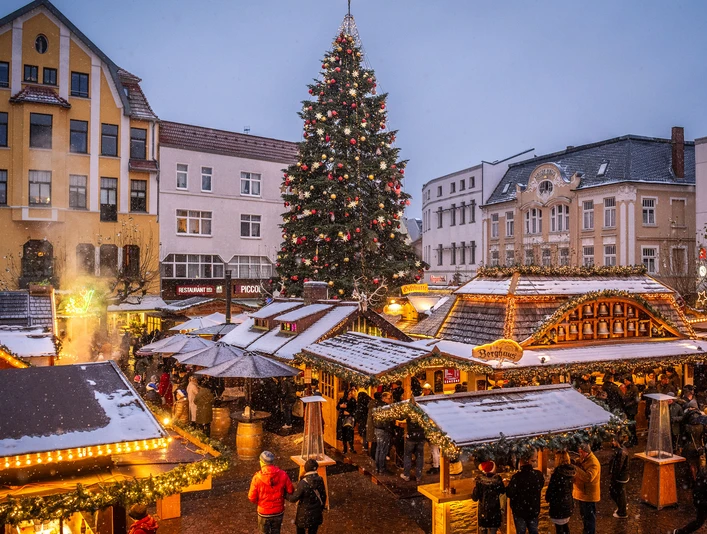 Weihnachtsmarkt mit leuchtendem Tannenbaum, geschmückten Ständen und Menschen in winterlicher Atmosphäre.