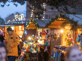 Weihnachtsmarkt mit beleuchteten Holzbuden unter Bäumen, Verkaufsstände voller bunter Waren und Besuchern.
