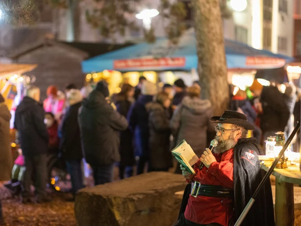 Ein Geschichtenerzähler fesselt Besucher eines Weihnachtsmarktes mit Lesung bei stimmungsvoller Beleuchtung.