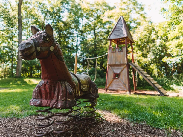 Spielplatz am Schlossberg Regenstauf Holzspielpferd auf Federn vor einem Kletterturm im grünen Außenbereich