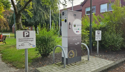 Ladestation E Auto Rathaus.jpg Eine Ladestation für Elektrofahrzeuge steht vor dem Rathaus, umgeben von Bäumen und Parkschildern.