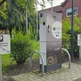 Ladestation E Auto Rathaus.jpg Eine Ladestation für Elektrofahrzeuge steht vor dem Rathaus, umgeben von Bäumen und Parkschildern.