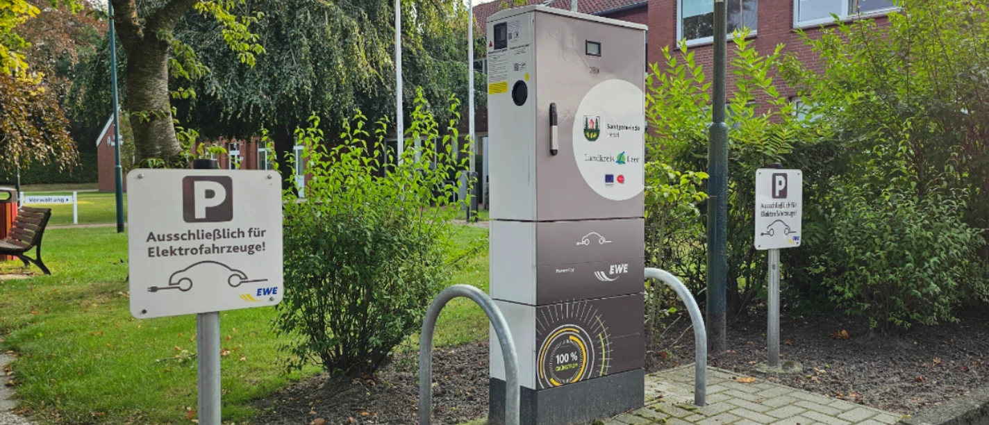 Ladestation E Auto Rathaus.jpg Eine Ladestation für Elektrofahrzeuge steht vor dem Rathaus, umgeben von Bäumen und Parkschildern.