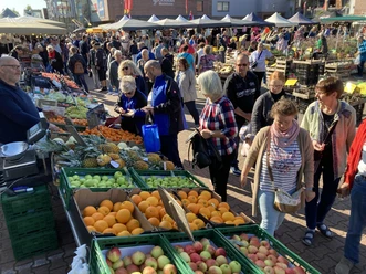 Hollandmarkt_an_der_Rhauderfehner_Textilwelt_Modehaus_BöckmannIMG_6157.jpg Markt mit vielen Menschen, die Obst und Gemüse kaufen. Im Vordergrund sind grüne Kisten mit Äpfeln, Orangen und Äpfeln. Im Hintergrund sind Stände mit weiteren Lebensmitteln.