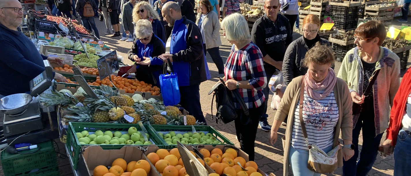 Hollandmarkt_an_der_Rhauderfehner_Textilwelt_Modehaus_BöckmannIMG_6157.jpg Markt mit vielen Menschen, die Obst und Gemüse kaufen. Im Vordergrund sind grüne Kisten mit Äpfeln, Orangen und Äpfeln. Im Hintergrund sind Stände mit weiteren Lebensmitteln.