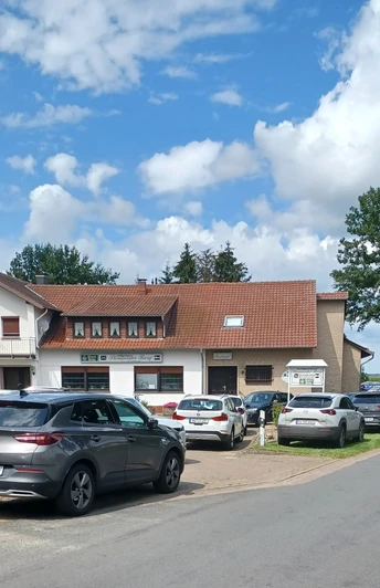 Parking lot with several cars in front of a building with a red roof and blue advertising, surrounded by trees.