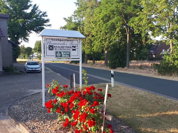 1000000158.jpg Hinweisschild des Landgasthauses Hermeshof Beck mit roten Blumen im Vordergrund und Straße daneben.