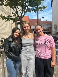 image_123650291.JPG Drei lächelnde junge Frauen stehen nebeneinander im Freien vor einem Gebäude mit Baum.