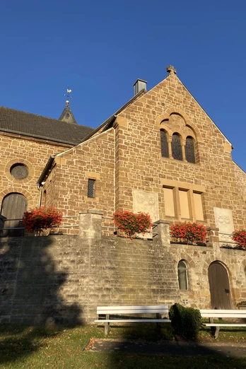 Kirche Bad Holzhausen.JPG Ein steinernes Kirchengebäude mit Blumen geschmückt, im Sonnenlicht einer klaren, blauen Tageszeit.