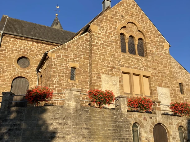 Kirche Bad Holzhausen.JPG Ein steinernes Kirchengebäude mit Blumen geschmückt, im Sonnenlicht einer klaren, blauen Tageszeit.