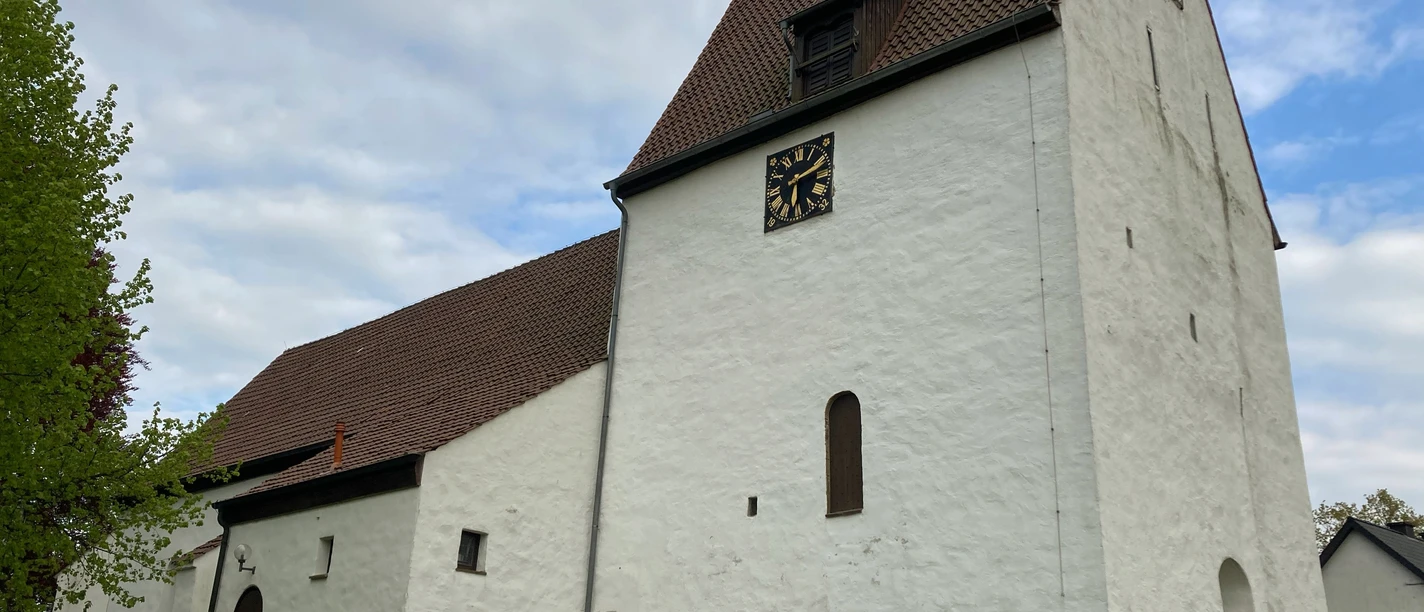 Weiße Kirche mit Satteldach und Turm, umgeben von einer niedrigen Steinmauer und Bäumen.