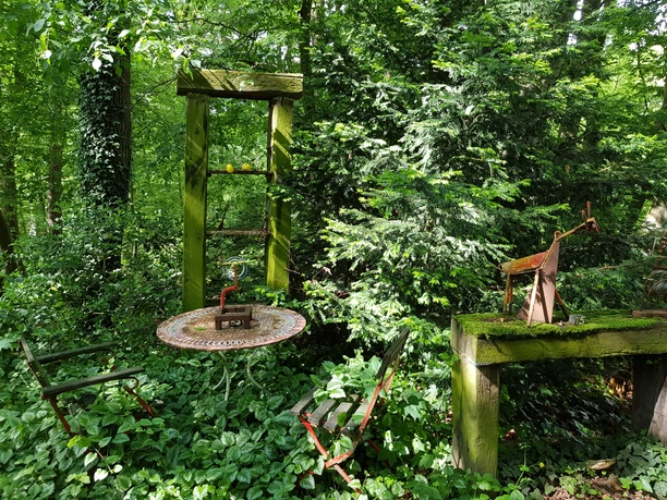Galerie Sattelgut Ein verwunschener Garten mit Moosbewuchs, Tisch und Stühlen, umgeben von üppigem Grün und Skulpturen.