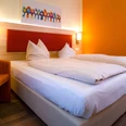Doppelzimmer im Hotel am Stadtpark Zwei helle Einzelbetten, orangefarbene Akzente im Zimmer, inklusive farbenfrohem Vogelbild an der Wand.