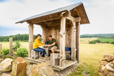 l-gde-kloster-falkenhagen-drehschutzh-tte-teutoburger-wald-tourismus-d-ketz-099-jpg.jpg Holzüberdachte Raststätte im Teutoburger Wald mit zwei Personen beim Picknick, umgeben von Natur.