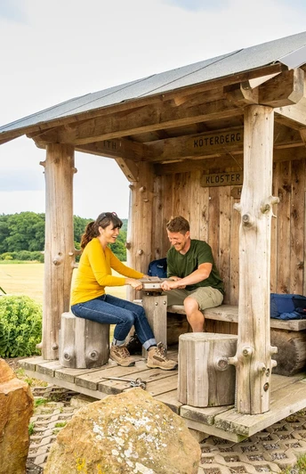 l-gde-kloster-falkenhagen-drehschutzh-tte-teutoburger-wald-tourismus-d-ketz-099-jpg.jpg Holzüberdachte Raststätte im Teutoburger Wald mit zwei Personen beim Picknick, umgeben von Natur.