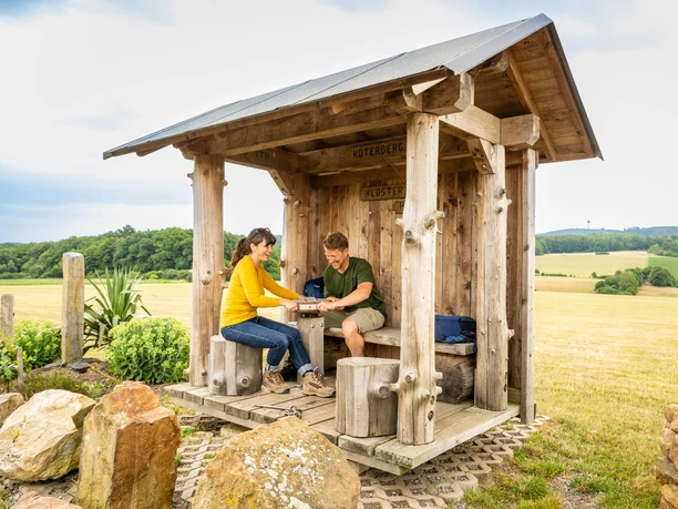 l-gde-kloster-falkenhagen-drehschutzh-tte-teutoburger-wald-tourismus-d-ketz-099-jpg.jpg Holzüberdachte Raststätte im Teutoburger Wald mit zwei Personen beim Picknick, umgeben von Natur.