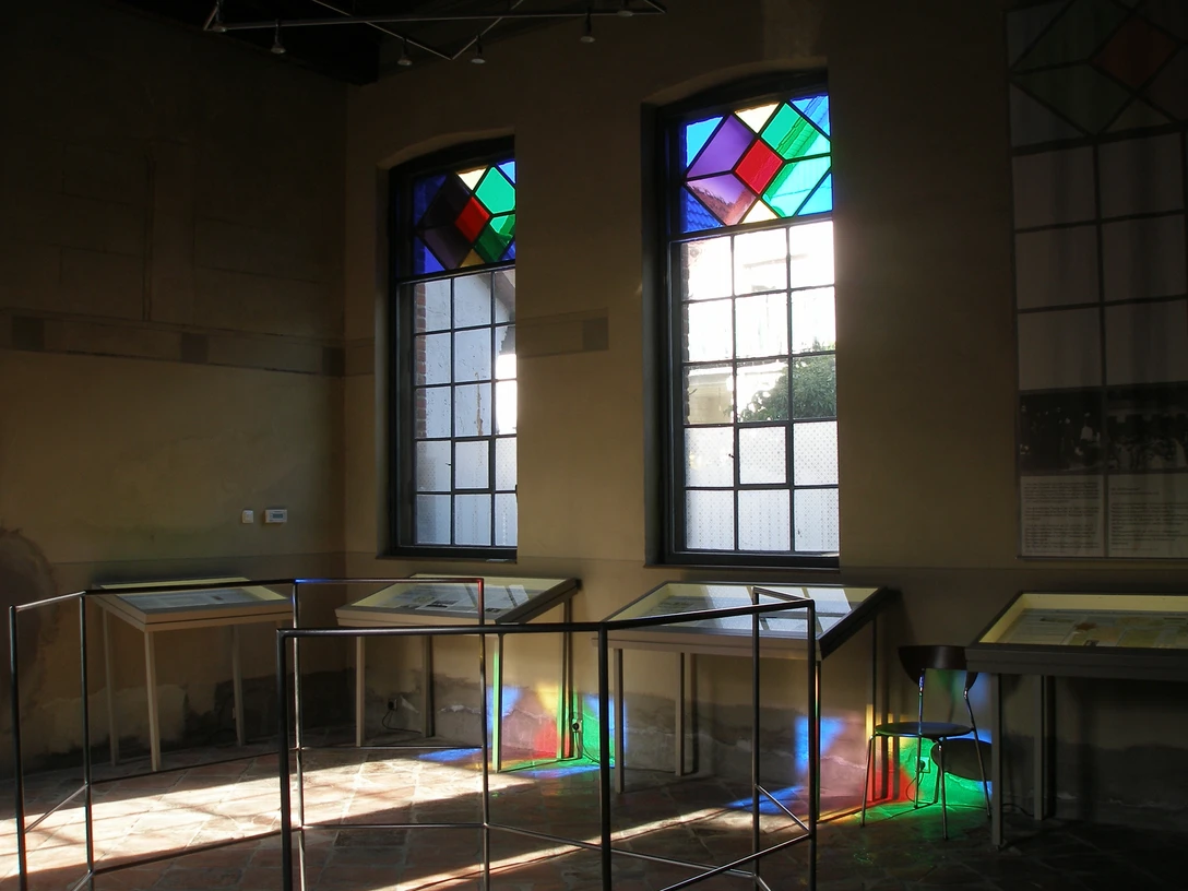 Stadt Petershagen_Petershagen_Evelyn Hotze_Alte Synagoge_Innenraum_Vitrinen_Spiegelung bunte Fenster_Sonne_quer.jpg Innenraum einer Synagoge mit Vitrinen und farbigen Glasfenstern, Sonnenstrahlen erzeugen bunte Reflexionen.