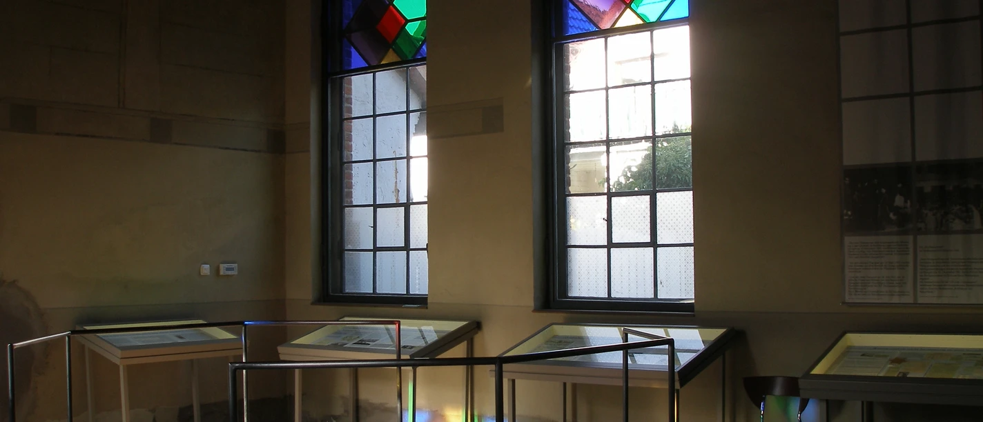 Stadt Petershagen_Petershagen_Evelyn Hotze_Alte Synagoge_Innenraum_Vitrinen_Spiegelung bunte Fenster_Sonne_quer.jpg Innenraum einer Synagoge mit Vitrinen und farbigen Glasfenstern, Sonnenstrahlen erzeugen bunte Reflexionen.