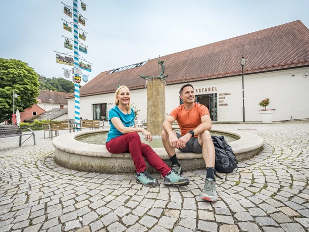 Rast in Pielenhofen Pärchen sitzt an einem Brunnen und macht Pause von ihrer Wanderung. Im Hintergrund send ein bayerischer Zunftbaum und das Klosterstadel Pielenhofen zu sehen.