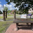 Pausenstation am Kreisel beim Rathaus Rhauderfehn Holztisch mit Bank auf gepflastertem Platz vor einer Infotafel und fünf Holzskulpturen in einem Park mit Bäumen und Gebäuden im Hintergrund