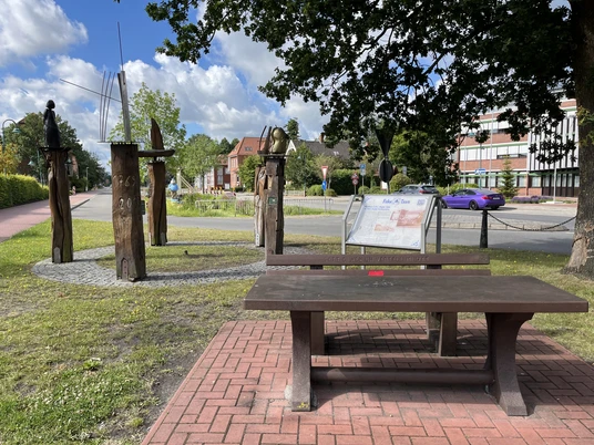 Pausenstation am Kreisel beim Rathaus Rhauderfehn Holztisch mit Bank auf gepflastertem Platz vor einer Infotafel und fünf Holzskulpturen in einem Park mit Bäumen und Gebäuden im Hintergrund