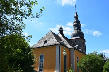 Evangelische Barockkirche Eckenhagen Barockkirche mit Zwiebeltürmen vor blauem Himmel, umgeben von grünen Bäumen und Laterne im Vordergrund.