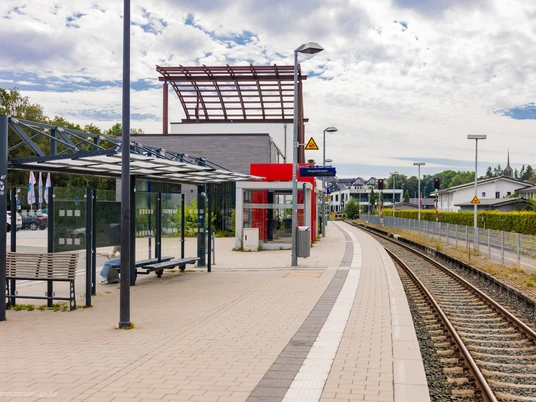 Bahnsteig mit Wartebereich, Bahngleisen und Bahnhof.