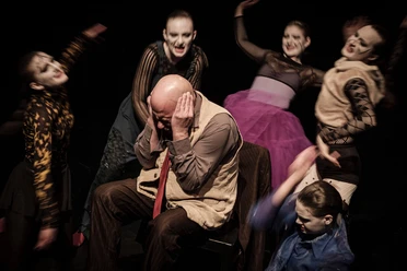 Theater der Keller Six masked actors dressed in imaginative costumes circle a man sitting in a chair with his ears covered. Their expressive gestures suggest an intense dramatic scene.