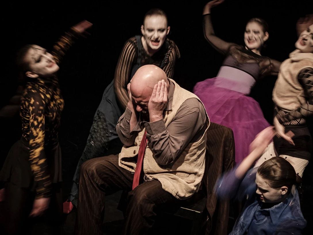 Theater der Keller Sechs in fantasievollen Kostümen gekleidete Schauspielende, maskiert, umkreisen einen Mann, der die Ohren zuhaltend auf einem Stuhl sitzt. Ihre ausdrucksstarken Gesten lassen eine intensive dramatische Szene vermuten.Six masked actors dressed in imaginative costumes circle a man sitting in a chair with his ears covered. Their expressive gestures suggest an intense dramatic scene.