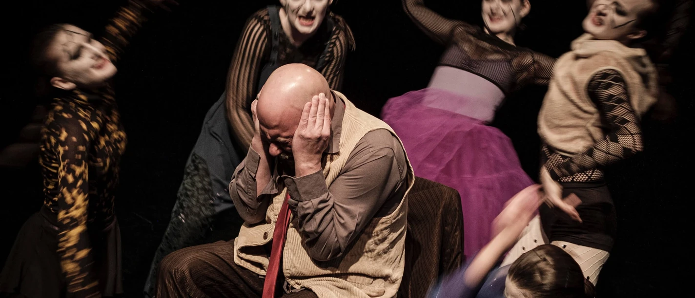 Theater der Keller Sechs in fantasievollen Kostümen gekleidete Schauspielende, maskiert, umkreisen einen Mann, der die Ohren zuhaltend auf einem Stuhl sitzt. Ihre ausdrucksstarken Gesten lassen eine intensive dramatische Szene vermuten.