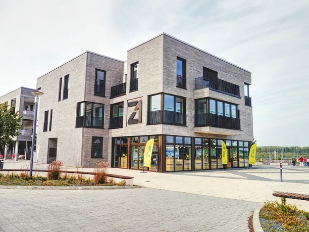 Zdjęcie przedstawia nowoczesne centrum informacji turystycznej Zwenkau z zewnątrz, za nim widać port na jeziorze Zwenkau.