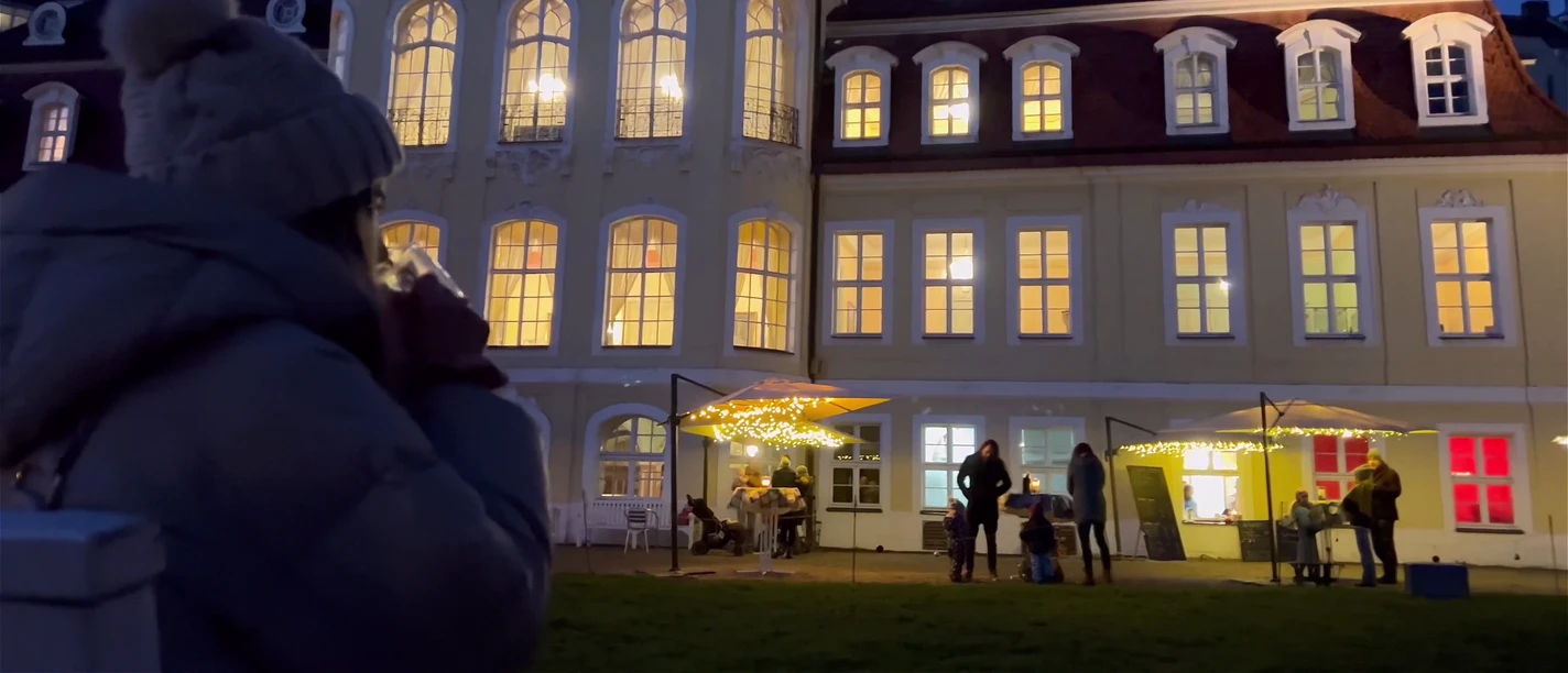 A woman with a hot drink in her hand looks at the illuminated Gohliser Schlösschen in the evening