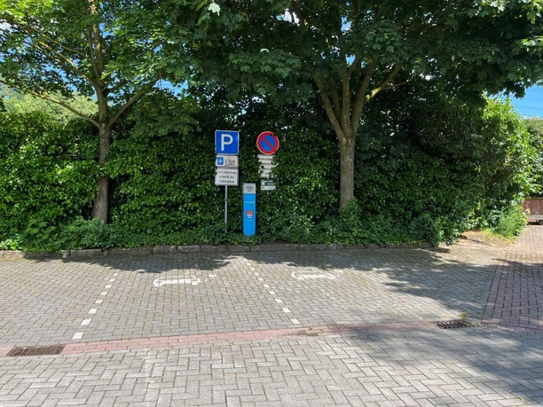 240528_WoMo-Stellpl, Dr-J-Stockmann-Str (Foto Gem. Salzbergen (2).jpg Parkplatz mit Stromsäule und zwei Parkschildern, umgeben von Bäumen und dichter Grünhecke.