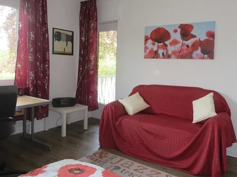 Helles Wohnzimmer mit rotem Sofa, weißen Kissen, Mohnblumenbild, Schreibtisch und Fenster mit Vorhang.