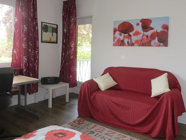 Helles Wohnzimmer mit rotem Sofa, weißen Kissen, Mohnblumenbild, Schreibtisch und Fenster mit Vorhang.