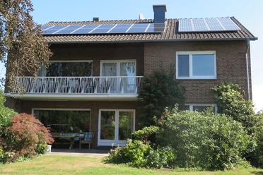 Zweistöckiges Ziegelhaus mit Solardach, großzügigem Balkon und gepflegtem Garten, Sonne scheint.