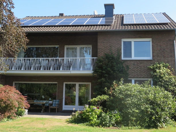 Zweistöckiges Ziegelhaus mit Solardach, großzügigem Balkon und gepflegtem Garten, Sonne scheint.