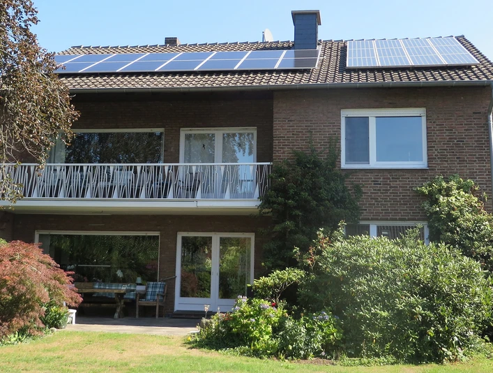 Zweistöckiges Ziegelhaus mit Solardach, großzügigem Balkon und gepflegtem Garten, Sonne scheint.