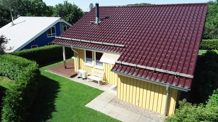 Ferienpark Extertal_Haus Lea Gelbes Ferienhaus mit rotem Dach umgeben von grüner Hecke und Rasenfläche, Sitzbank auf Terrasse.